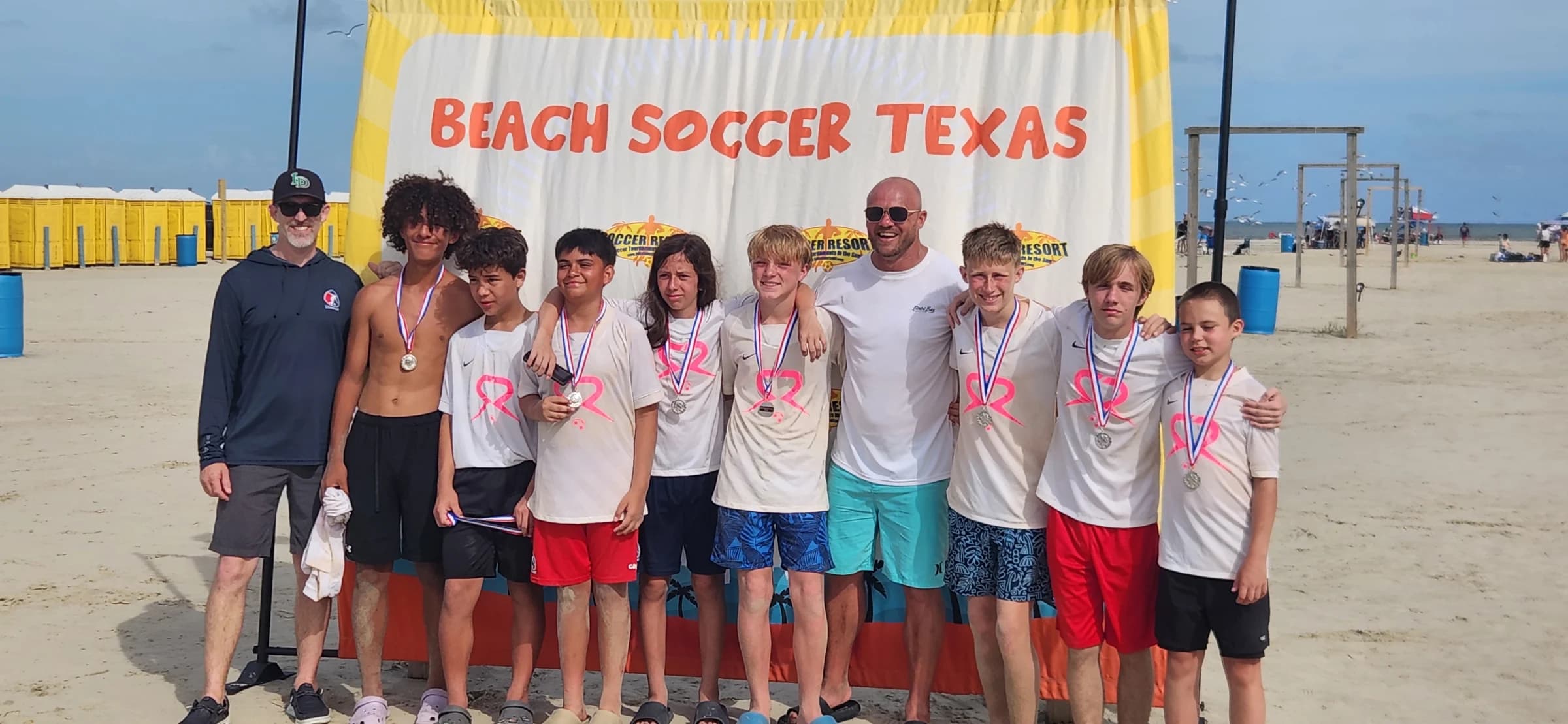 SOLO Soccer players and coaches wearing tournament medals beneath the Beach Soccer Texas banner in Galveston